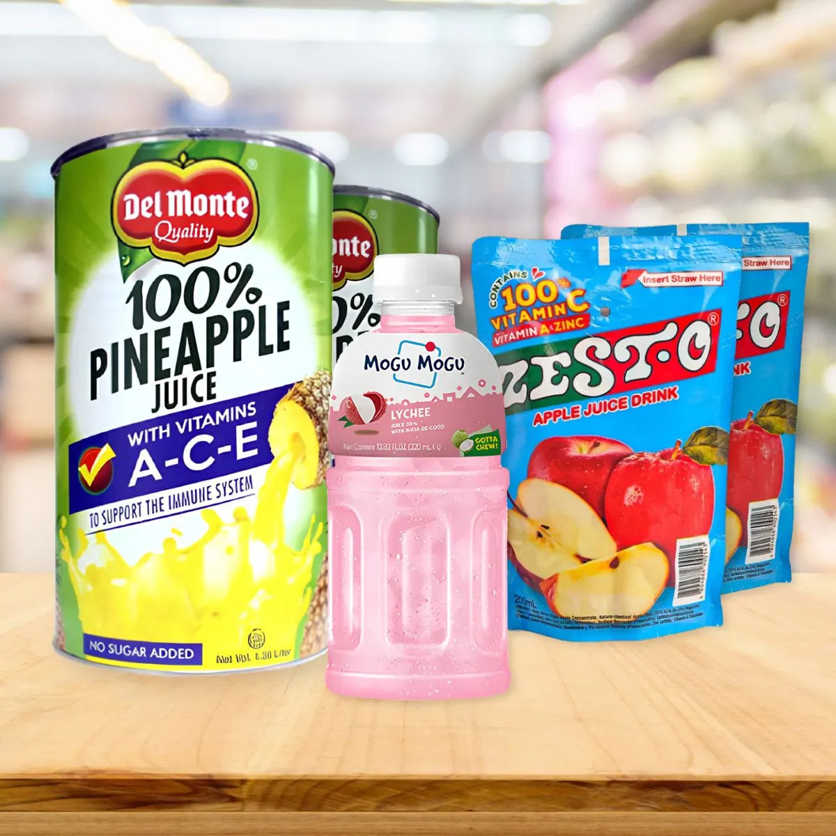 Juice drink display featuring a Del Monte Pineapple Juice can on the left, a Mogu Mogu Lychee bottle in the center, and two Zesto Apple pouches on wood.